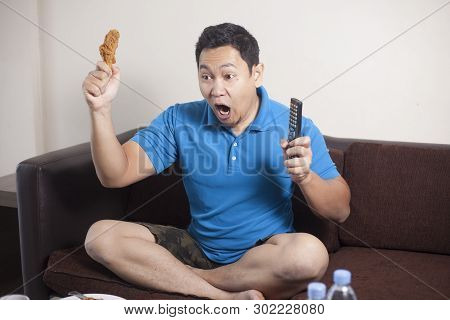 Portrait Of Young Funny Asian Man Watching Football Or Soccer Game On Tv On Cluch With Some Food