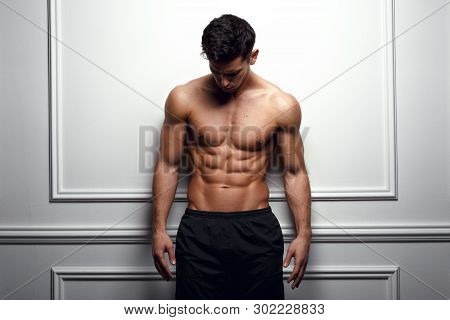 Athlete, Muscular Man At The White Wall Poses Shirtless, Showing Six Pack Abs, White Background.