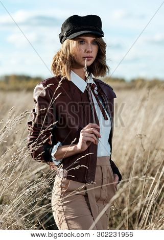 Portrait Of Attractive Female Model In Leather Jacket And Fashionable Cap Looking At Camera Against 