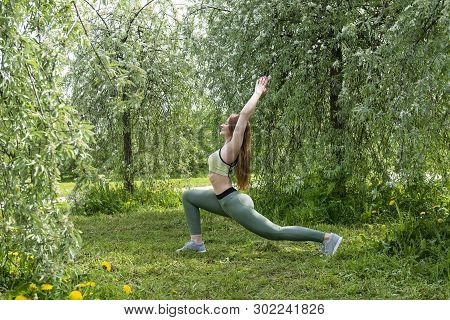 1 White Sports Girl Performs The Yoga Pose Warrior In Nature Among Trees And Greenery, Training Outd