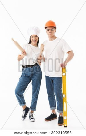 Full length portrait of a young attractive man architect and woman chef standing isolated over white background, holding building level and rolling pin