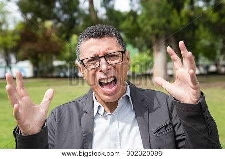 Indignant Middle-aged Man Screaming And Raising Hands In Park. Guy Wearing Casual Clothes And Lookin