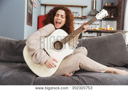 Photo of pretty young woman wearing house clothes playing acoustic guitar while sitting on sofa in apartment