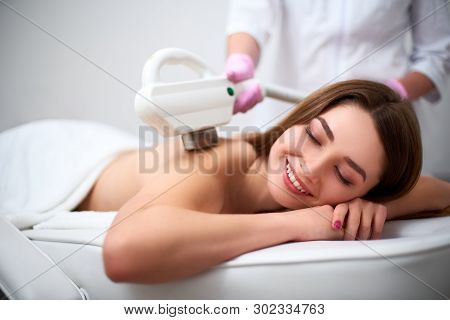 Young Pretty Smiling Woman Back Laser Epilation In Beauty Salon. Cosmetologist Doing Depilation Trea
