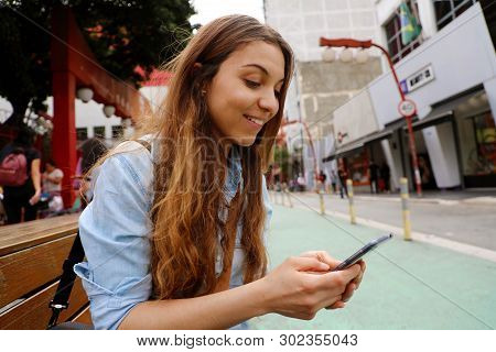 Beautiful Student Girl Sitting On Street Bench Messaging With Mobile Phone In Sao Paulo City, Brazil