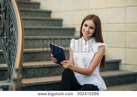 Young And Attractive Female Sittind On The Stairs And Use Tablet. Woman Play Online Games On Tablet