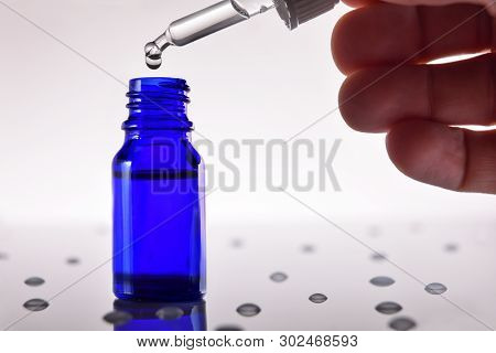 Hand Putting Drops From A Dropper In A Blue Jar Full Of Liquid On Glass Table Full Of Drops. Horizon