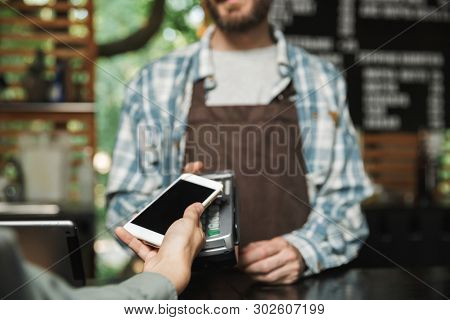 Portrait of caucasian barista man wearing apron holding consumer terminal for paypass payment while working in street cafe or coffeehouse outdoor