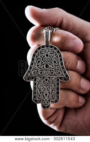 closeup the hand of a young man holding an old hamsa amulet, also known of the hand of fatima or the hand of mary, against a black background