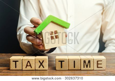 Wooden Blocks With The Word Tax Time And A Miniature House In The Hands Of A Businessman. The Concep