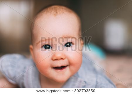 Cute, Caucasian, Smiling Baby With Open Mouth In The Grey Bodysuit Looks Slyly Into The Lens In The 