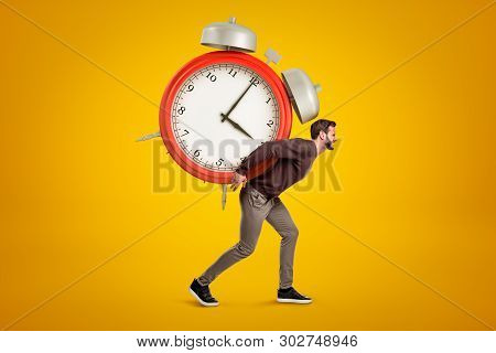 Side View Of Young Handsome Man Carrying Huge Alarm Clock On His Back On Yellow Gradient Background.