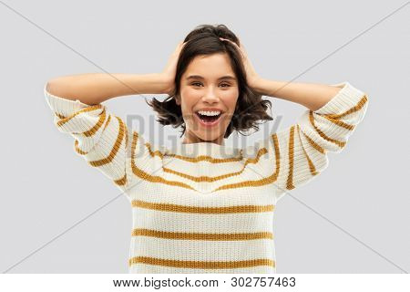 emotion, expression and people concept - happy smiling young woman in striped pullover holding to her head over grey background