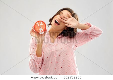 people and bedtime concept - sleepy young woman in pajama with alarm clock showing half past six stretching and yawning over grey background