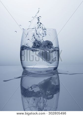 Ice cubes fall into a pot-bellied glass of clean, pure drinking water. Splashing in water, close-up shot with reflection on white background