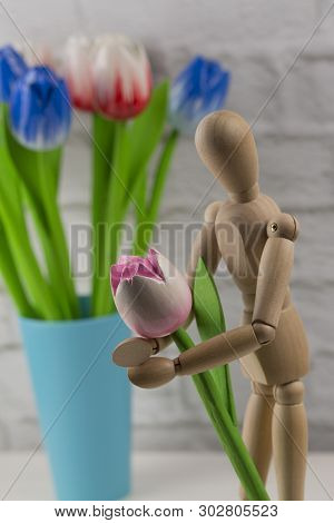 Wooden man holding a wooden pink flower. A man with a flower tulips in his hands, a concept - waiting for a date with his beloved. Artificial wooden multi-colored flowers as a symbol of love