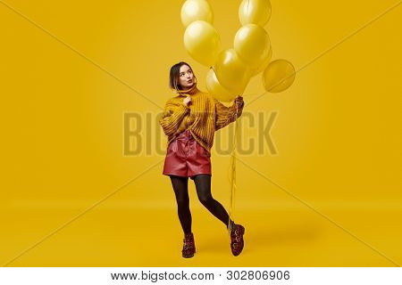 Modern Young Lady In Stylish Outfit Holding Bunch Of Vivid Balloons While Standing Against Yellow Ba