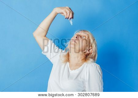 An Old Woman Is Dripping Herself A Drop In Her Eyes Against A Blue Background. Concept Of Eye Treatm