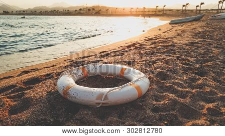 Closeup Toned Image Of Life Saving Ring Lying On The Sea Baech Sand. Beutiful Sunset Over The Ocean
