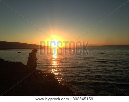 Silhouette Of A Girl At Sunset On Lake Baikal. The Bright Sun Hangs Over The Horizon And Beautifully