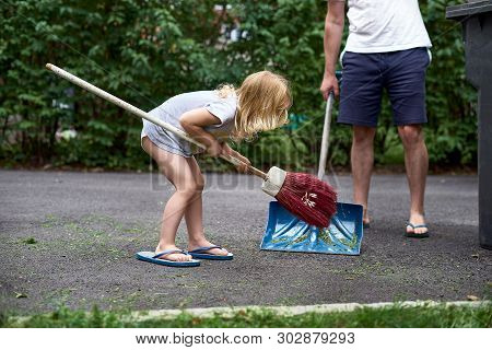 Little Girl Helps Her Parents Collect Grass In The Yard. Seasonal Gardening. Cleaning The Yard