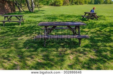 Wooden Picnic Tables On Grass Parkland In The Shade.  Lady Sitting At One Of The Tables.