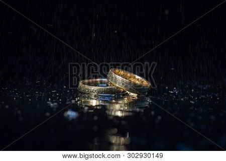 Wedding Rings Lying On Dark Surface Shining With Light Close Up Macro. Water Splashes