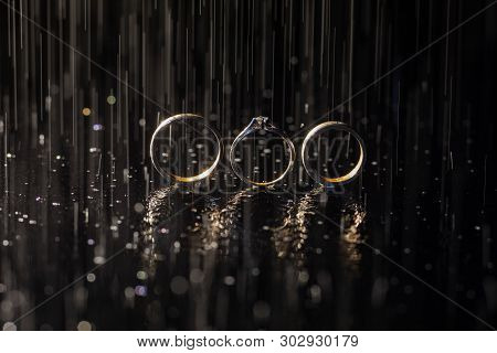 Wedding And Engagement Rings Lying On Dark Surface Shining With Light Close Up Macro. Water Splashes