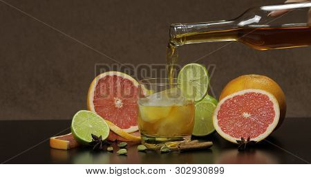 Pour Liquor From A Bottle Into A Glass Cup With Ice Cubes. Fresh Fruits In The Background. Lime Grap