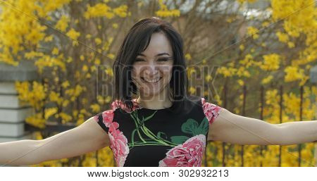 Attractive Young Woman In A Dress With Flowers Making Funny Faces Near Fence. Cute Girl With Black H