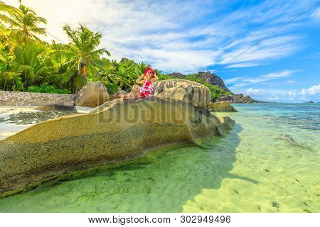 La Digue, Seychelle. Lifestyle Tourist Woman With Red Hat Above A Huge Granite Boulder At Anse Sourc