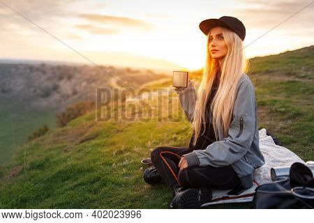 Portrait Of Young Blonde Girl With Black Cap, Holding Steel Mug With Hot Tea On Background Of Sunset