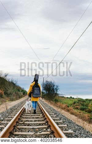 Unrecognizable Musician In A Yellow Coat Walking Her Dog Along The Train Tracks With The Guitar Load