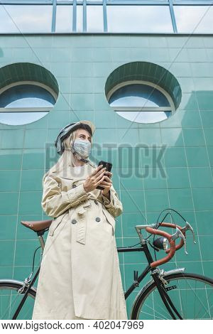 Woman in protective maskin, in helmet using her smartphone device. Female go to work on his sport retro bike. Girl standing on green background.