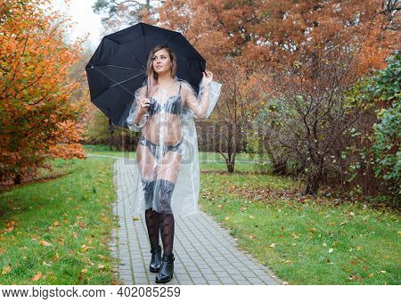 Young Woman In Black Lingerie, Stockings And A Transparent Raincoat With Umbrella Standing On The Al