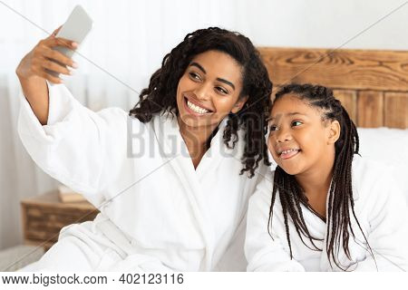 Happy Black Mother And Daughter In Bathrobes Taking Selfie On Bed, Capturing Self-portrait On Smartp