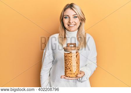 Young caucasian woman holding jar of macaroni pasta looking positive and happy standing and smiling with a confident smile showing teeth 