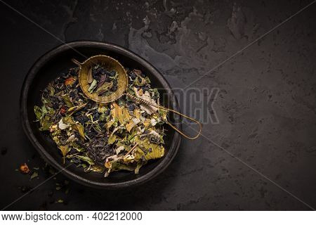 Mixture of teas, dried organic tea herbs and fruits in bowl with tea strainer on black background