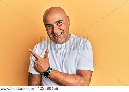 Mature middle east man wearing casual white tshirt cheerful with a smile of face pointing with hand and finger up to the side with happy and natural expression on face 