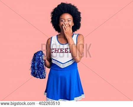 Young african american woman wearing cheerleader uniform holding pompom laughing and embarrassed giggle covering mouth with hands, gossip and scandal concept 
