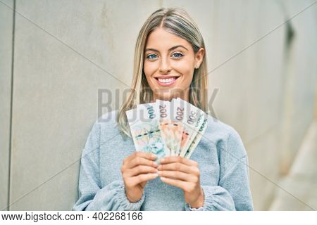 Young blonde girl smiling happy holding czech koruna banknotes at the city.