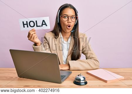 Beautiful hispanic woman wearing operator headset holding hola greenting scared and amazed with open mouth for surprise, disbelief face 