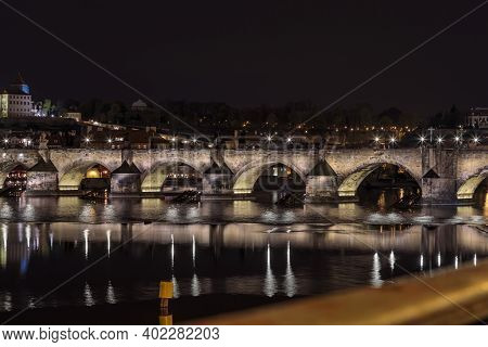 Charles Bridge In Prague In The Evening With Colorful Lights From Lanterns. In The River The Reflect