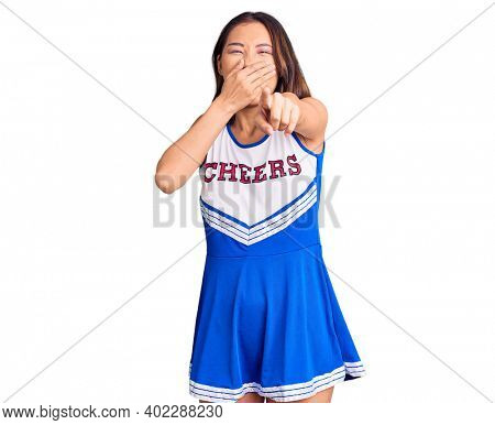 Young beautiful chinese girl wearing cheerleader uniform laughing at you, pointing finger to the camera with hand over mouth, shame expression 