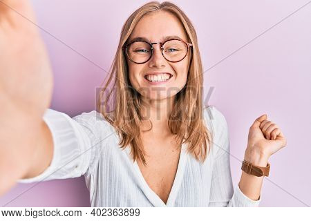 Beautiful blonde woman taking a selfie photo wearing glasses screaming proud, celebrating victory and success very excited with raised arm 