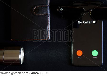 A Smartphone With An Unidentified Call Lies On A Dark Surface Next To Black Sunglasses, A Notebook A