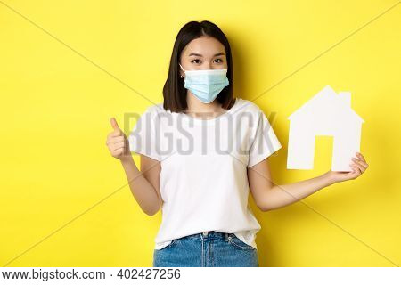 Covid-19, Pandemic And Real Estate Concept. Pretty Asian Woman Smiling In Medical Mask, Showing Pape
