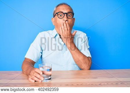 Senior handsome man with gray hair drinking glass of water sitting on the table covering mouth with hand, shocked and afraid for mistake. surprised expression 