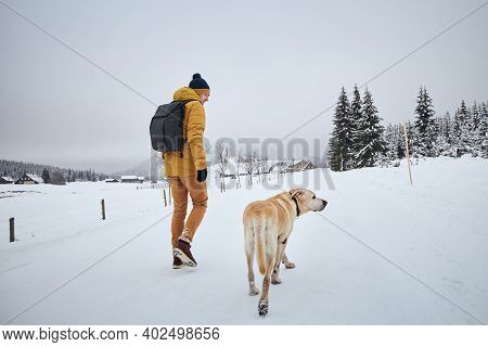 Rear View Of Young Man With Dog In Winter. Pet Owner With His Labrador Retriever Walking Against Old