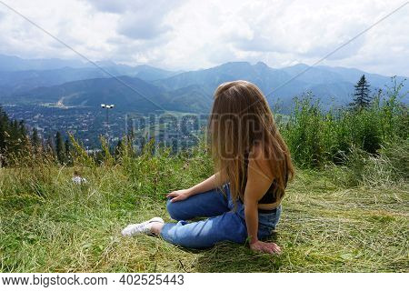 A Girl With Dark Hair, In Jeans And A Black T-shirt, Sits On A Lawn And Looks At The Mountains. View